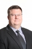 Man in dark suit jacket, wearing light checkered shirt, dark tie and glasses.